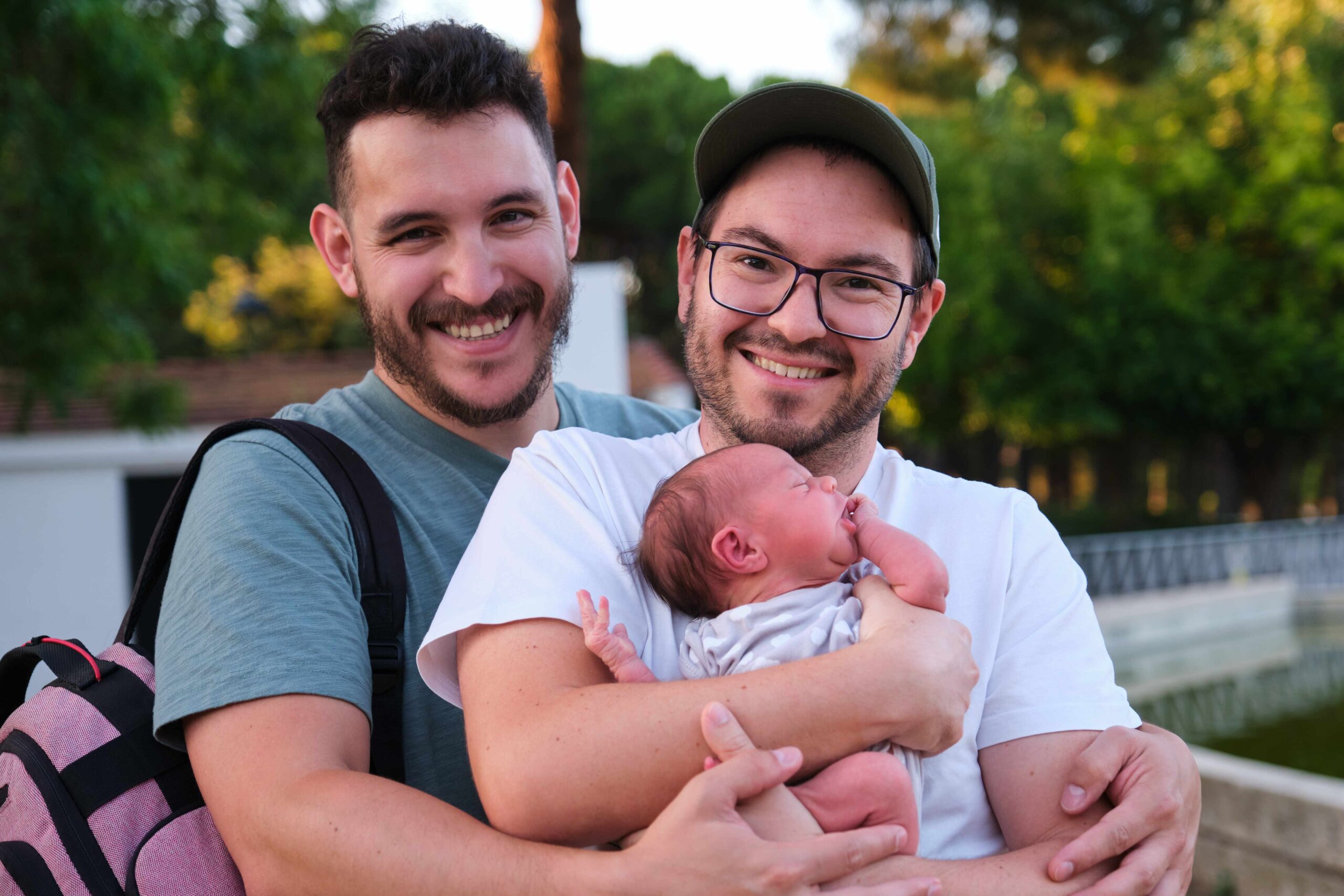 Gay couple with their newborn baby in a park smiling and looking at camera.